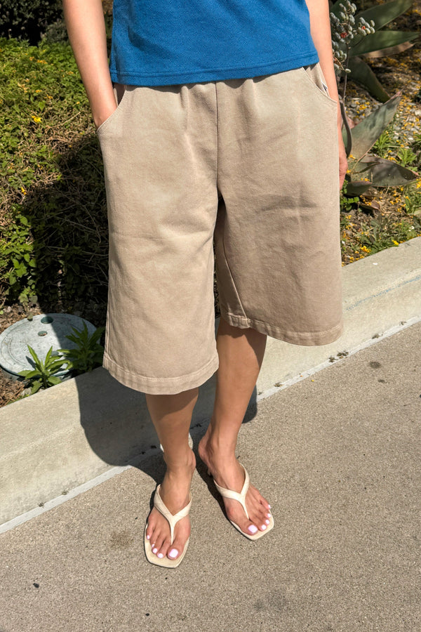 Person wearing beige shorts and a blue top on a sidewalk with plants in the background