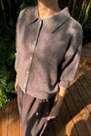 A person standing on a wood deck wearing brown pants and a brown cashmere collared button up cardigan.