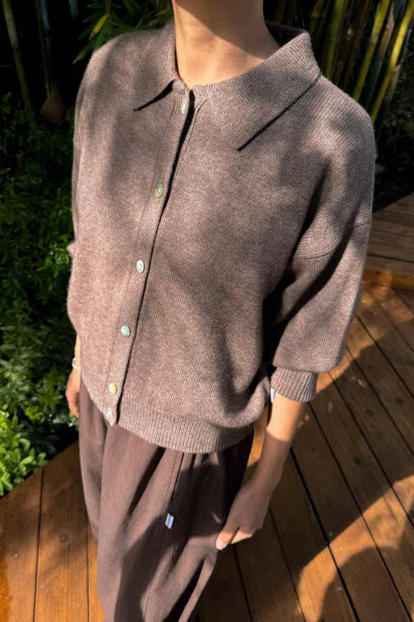 A person standing on a wood deck wearing brown pants and a brown cashmere collared button up cardigan.