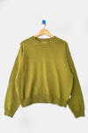 A lightweight cotton knit crew sweater in a yellow-green shade hanging on a wood hanger against a white wall.
