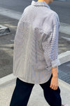 View of a person walking away from the camera down a sidewalk wearing dark pants and a blue and white striped overshirt with the sleeves rolled up.