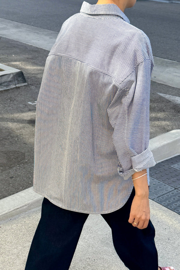 View of a person walking away from the camera down a sidewalk wearing dark pants and a blue and white striped overshirt with the sleeves rolled up.