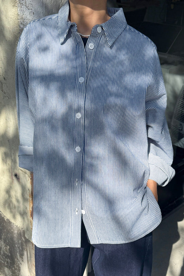 A person wearing a blue and white striped button up shirt with a pair of dark pants standing against a concrete wall under dappled shadows.