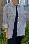 A person wearing a blue and white striped shirt styled open over a dark shirt and pants with the sleeves rolled up. They are standing outside with plants and a tree in the background.