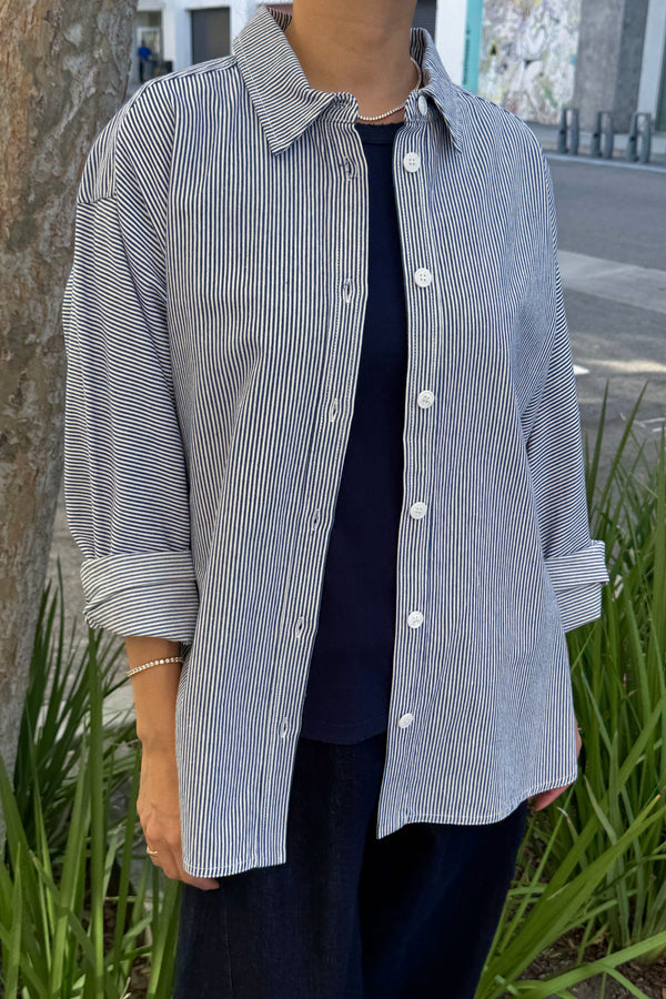 A person wearing a blue and white striped shirt styled open over a dark shirt and pants with the sleeves rolled up. They are standing outside with plants and a tree in the background.