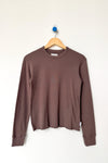 A brown long-sleeve crewneck tee hanging on a wooden hanger against a plain white wall.