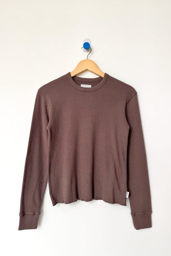 A brown long-sleeve crewneck tee hanging on a wooden hanger against a plain white wall.