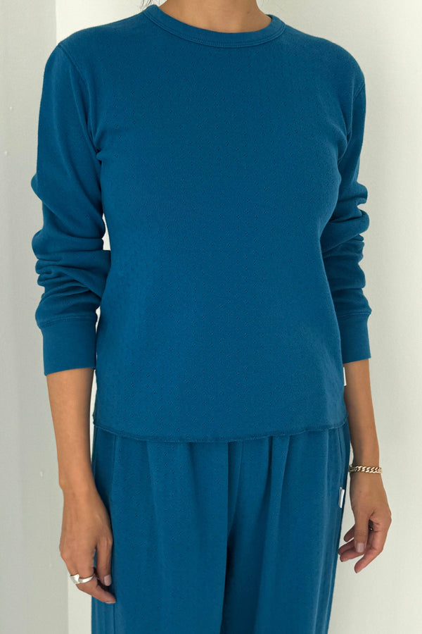 A person wearing a deep teal blue long sleeve knit tee with the sleeves pushed up over a pair of matching pants against a white wall.