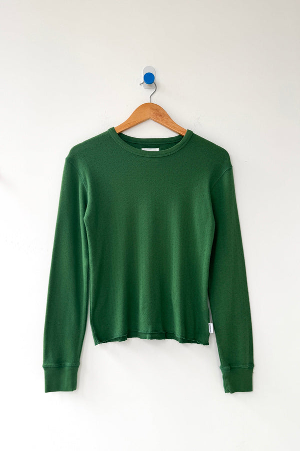 A moss green pointelle knit long sleeve tee hanging on a wood hanger against a white wall.