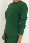 A person wearing a rich green knit long sleeve tee with the sleeves pushed up over a matching pair of pants standing against a white wall.