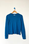 A dark teal blue lightweight knit long sleeve top hanging on a wood hanger against a white wall.