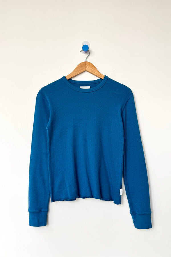 A dark teal blue lightweight knit long sleeve top hanging on a wood hanger against a white wall.