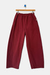 A pair of oxblood red barrel leg heavyweight cotton pants on a wood hanger against a white wall.