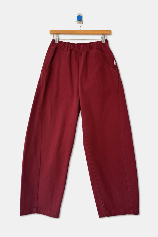 A pair of oxblood red barrel leg heavyweight cotton pants on a wood hanger against a white wall.