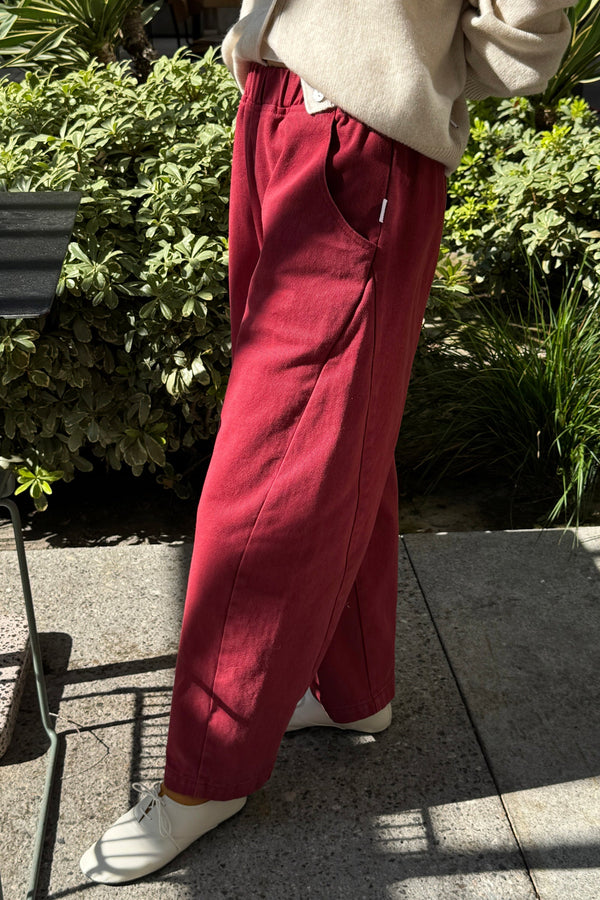 A person wearing a cream cardigan and oxblood red barrel leg pants with cream shoes, standing outside on a sidewalk near a garden.
