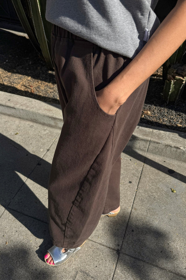 A person wearing dark brown wide-leg pants with side pockets, paired with a gray top and silver sandals, standing outdoors on a paved walkway.