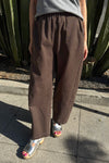 A person wearing loose brown wide-leg pants with a gray top and metallic silver sandals, standing outdoors on a concrete sidewalk in front of a cactus plant.
