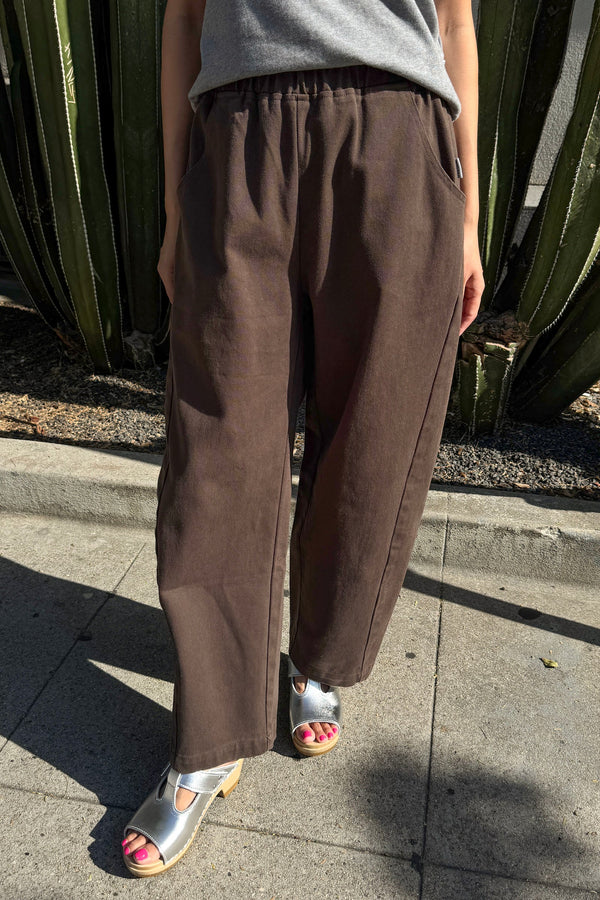 A person wearing loose brown wide-leg pants with a gray top and metallic silver sandals, standing outdoors on a concrete sidewalk in front of a cactus plant.