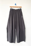 Dark gray wide-leg cotton knit pants with an elastic waistband, hanging from a wooden hanger against a plain white wall.