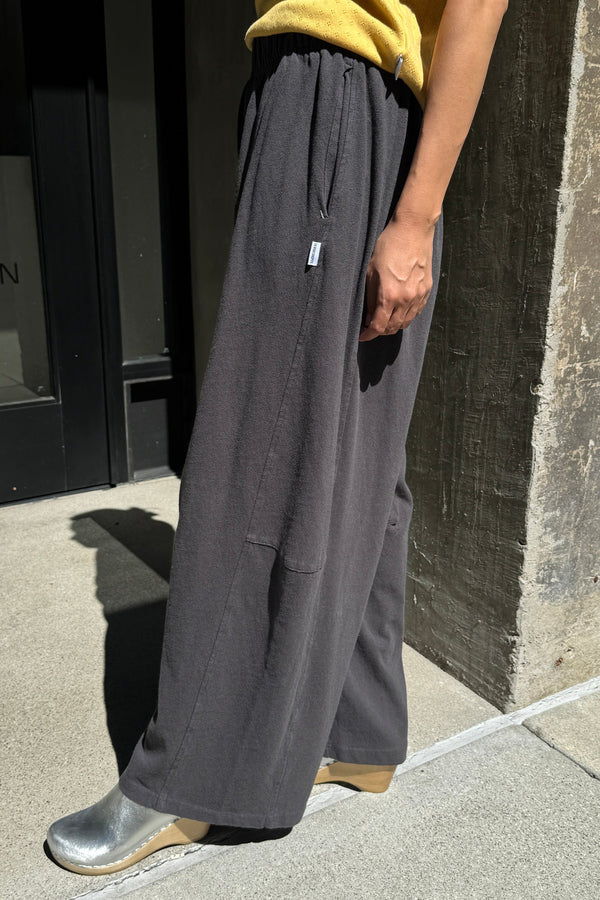 A person wearing loose, wide-leg dark gray pants with a yellow top and metallic silver boots, standing outdoors on a concrete surface next to a dark doorway.