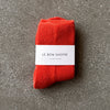 A pair of plush socks in a vibrant orange-red color with their white paper 'Le Bon Shoppe' packaging photographed on a grey backdrop. 