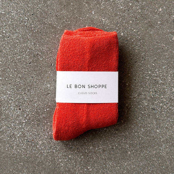A pair of plush socks in a vibrant orange-red color with their white paper 'Le Bon Shoppe' packaging photographed on a grey backdrop. 