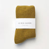 A pair of plush olive green socks in their white paper 'Le Bon Shoppe' packaging on a white surface.