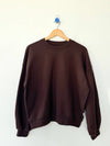 A dark brown crewneck sweatshirt hanging on a wooden hanger against a plain white wall.