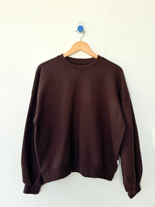 A dark brown crewneck sweatshirt hanging on a wooden hanger against a plain white wall.