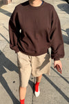 Person wearing a brown sweatshirt and beige shorts on a sidewalk.