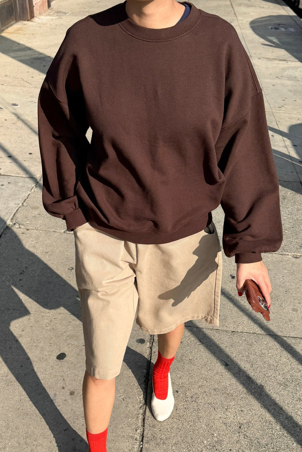 Person wearing a brown sweatshirt and beige shorts on a sidewalk.