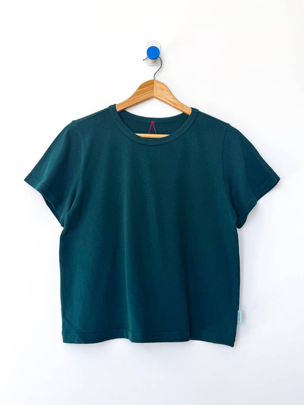 A dark forest green tee on a wood hanger against a white wall.
