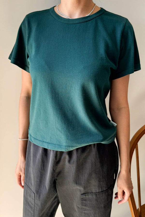 A person wearing a dark forest green short-sleeve crewneck t-shirt and dark gray pants, standing indoors against a light beige wall.