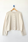 A cream-colored long-sleeve cropped snap closure cardigan hanging on a wooden hanger against a plain white wall.