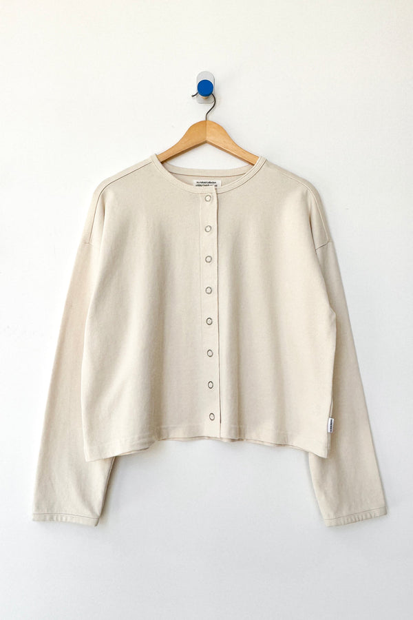 A cream-colored long-sleeve cropped snap closure cardigan hanging on a wooden hanger against a plain white wall.