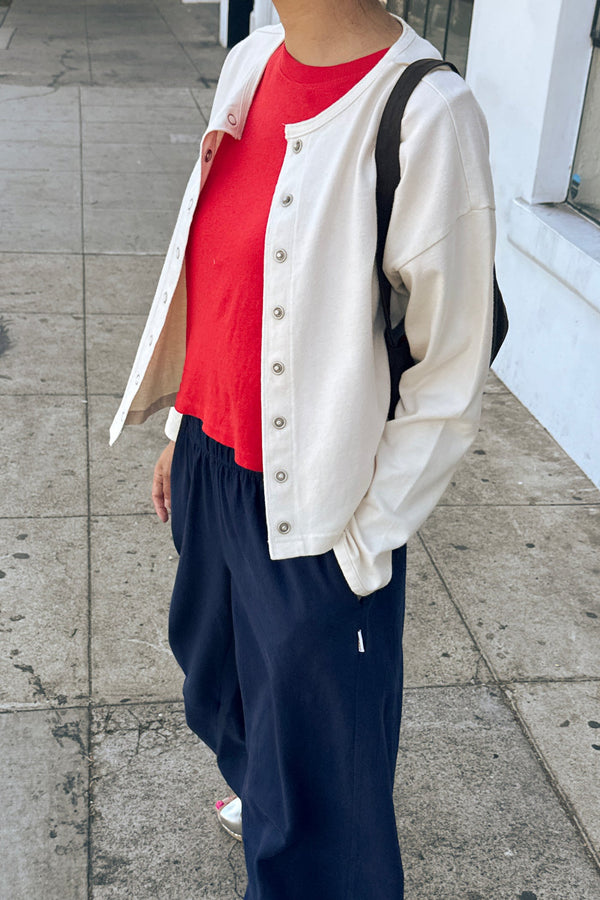 A person standing on a sidewalk wearing a cream-colored snap closure cardigan over a bright red t-shirt, paired with dark navy wide-leg pants and a black shoulder bag.