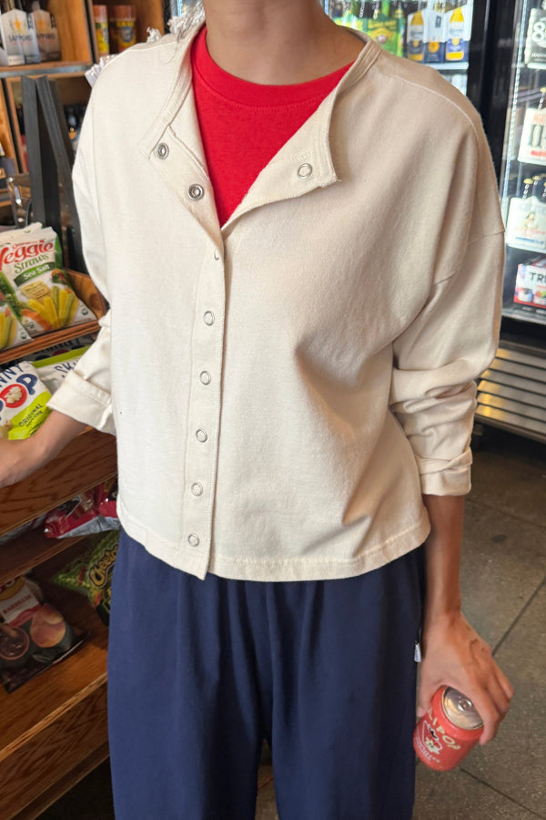 A person wearing a cream-colored, long-sleeve snap closure cardigan over a bright red t-shirt and navy blue pants, standing in a convenience store.