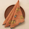 An orange-brown folded cloth napkin with yellow and green floral print and dark navy stitched edges, displayed on a round brown plate against a warm beige background.