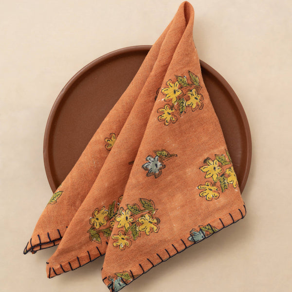 An orange-brown folded cloth napkin with yellow and green floral print and dark navy stitched edges, displayed on a round brown plate against a warm beige background.