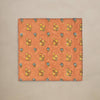 A square orange linen napkin with a repeating floral pattern in yellow, blue, and green, laid flat against a neutral beige background.