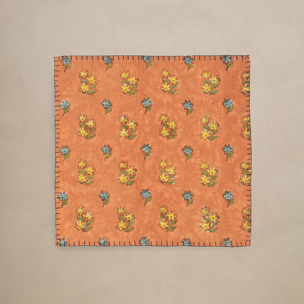 A square orange linen napkin with a repeating floral pattern in yellow, blue, and green, laid flat against a neutral beige background.