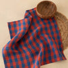 A red and navy blue plaid checkered cloth napkin styled on a beige surface with a wooden bowl and a woven placemat.