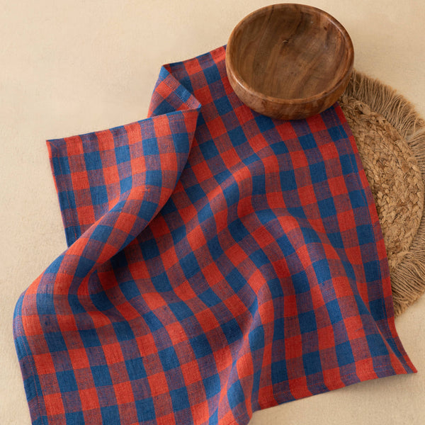 A red and navy blue plaid checkered cloth napkin styled on a beige surface with a wooden bowl and a woven placemat.