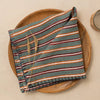 A linen napkin in rich green, red, orange, and white stripes with dark blanket stitch trim folded against a wooden plate on a neutral table.