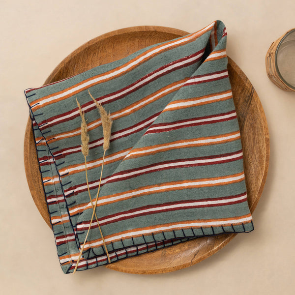 A linen napkin in rich green, red, orange, and white stripes with dark blanket stitch trim folded against a wooden plate on a neutral table.