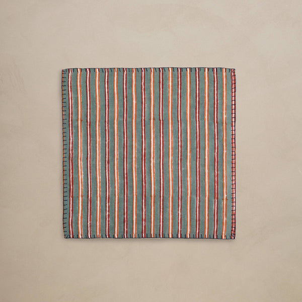 A folded linen napkin featuring a rich striped pattern in dark green, orange, red, and white, with dark red blanket stitching trim against a neutral backdrop.