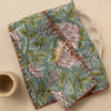 A stack of two folded linen napkins featuring a rich blue, green, white, and red floral design with red stitching on the edges. They lay against a ceramic plate next to a ceramic cup against a neutral backdrop.