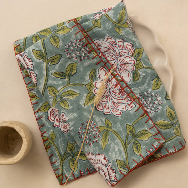 A stack of two folded linen napkins featuring a rich blue, green, white, and red floral design with red stitching on the edges. They lay against a ceramic plate next to a ceramic cup against a neutral backdrop.