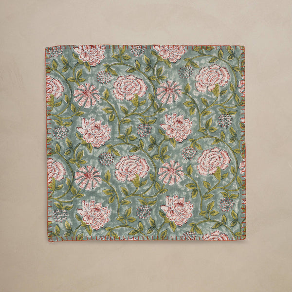 A linen table napkin in a rich blue, green, red, and white floral design with red blanket stitching along the edges, set against a neutral backdrop.