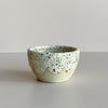 A mini ceramic bowl in a natural clay color featuring an off-white and brown speckled glaze with a wavy edge, sat against a neutral backdrop.